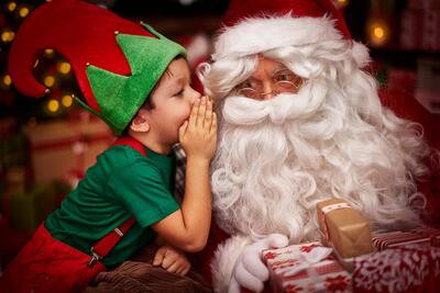 Ein Kind, als Elf verkleidet, flüstert dem Weihnachtsmann was ins Ohr.