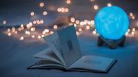 Bild vergrößern: Eine hellblaue Kristallkugel auf einer hölzernen Fassung steht hinter einem aufgeblätterten, schwarzweißen Bilderbuch. Im Hintergrund leuchten verschwommen Lichter.